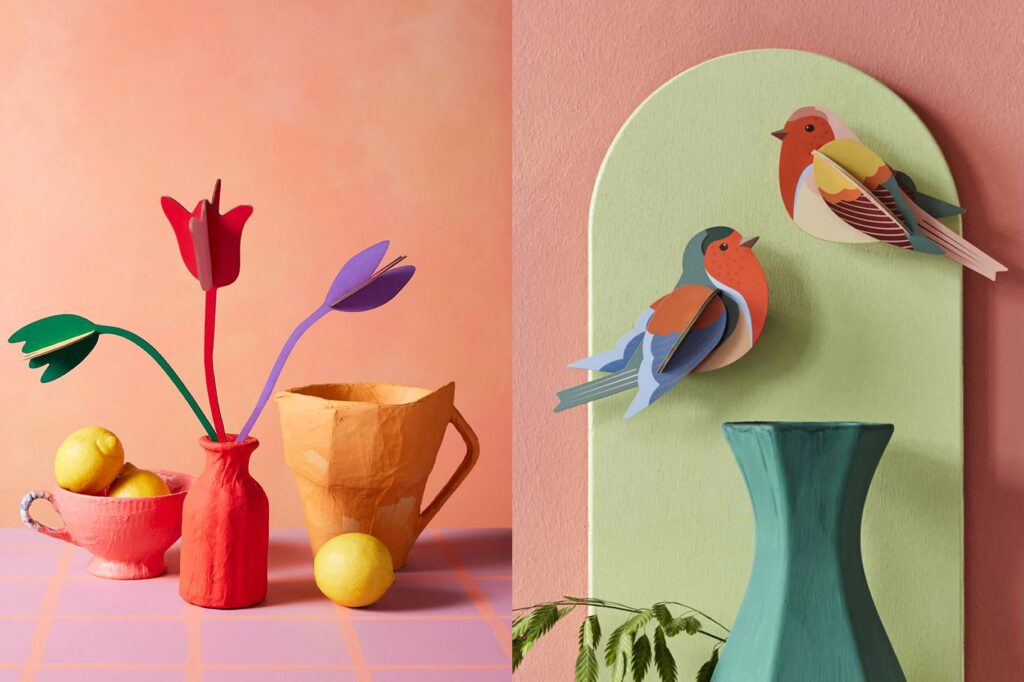 Two styled product photos side by side. Left: colourful paper tulips in red and purple arranged in a terracotta-toned paper vase, alongside a pink teacup with lemons and an orange paper mug, set against a peach background with a pink tile surface. Right: two 3D paper birds in blue and rust tones perched on a sage green arch-shaped wall decoration, beside a teal ceramic vase against a dusty pink wall.