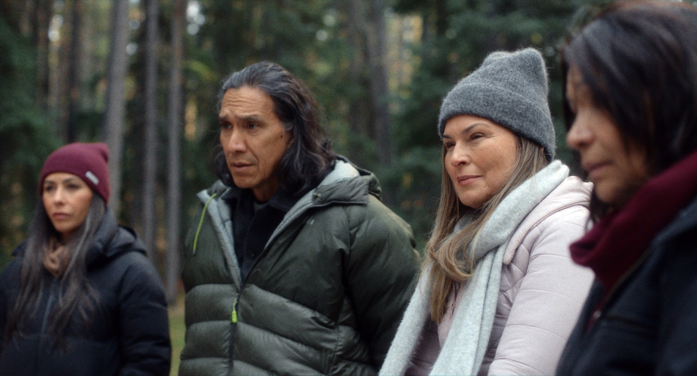 Four people in winter jackets and hats stand together in a forest, looking ahead with focused, serious expressions.
