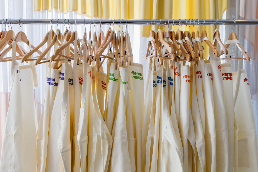 A row of cream-coloured canvas painting aprons hanging on wooden hangers along a metal rack. Each apron features the Remai Modern logo printed in different brand colours — including red, blue, green, orange, and yellow — with soft natural light filtering through sheer curtains behind them.