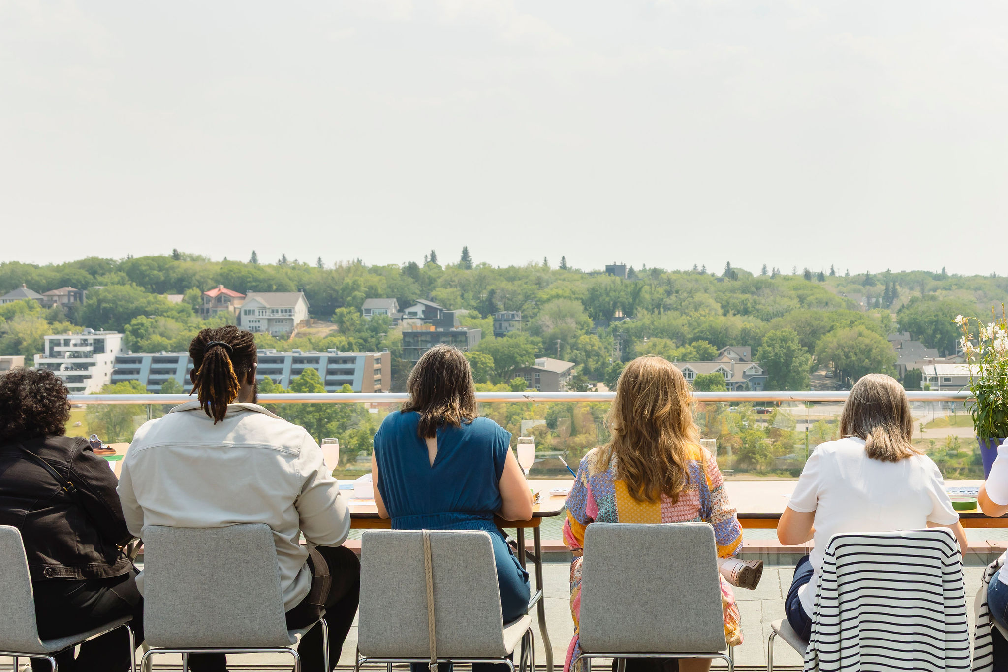 Field Study Rooftop Website Image Six participants sit with their backs to the camera at a long table on Remai Modern's rooftop patio, painting watercolours while looking out over a panoramic view of Saskatoon's South Saskatchewan River valley and tree-covered riverbank neighbourhoods on a sunny day.
