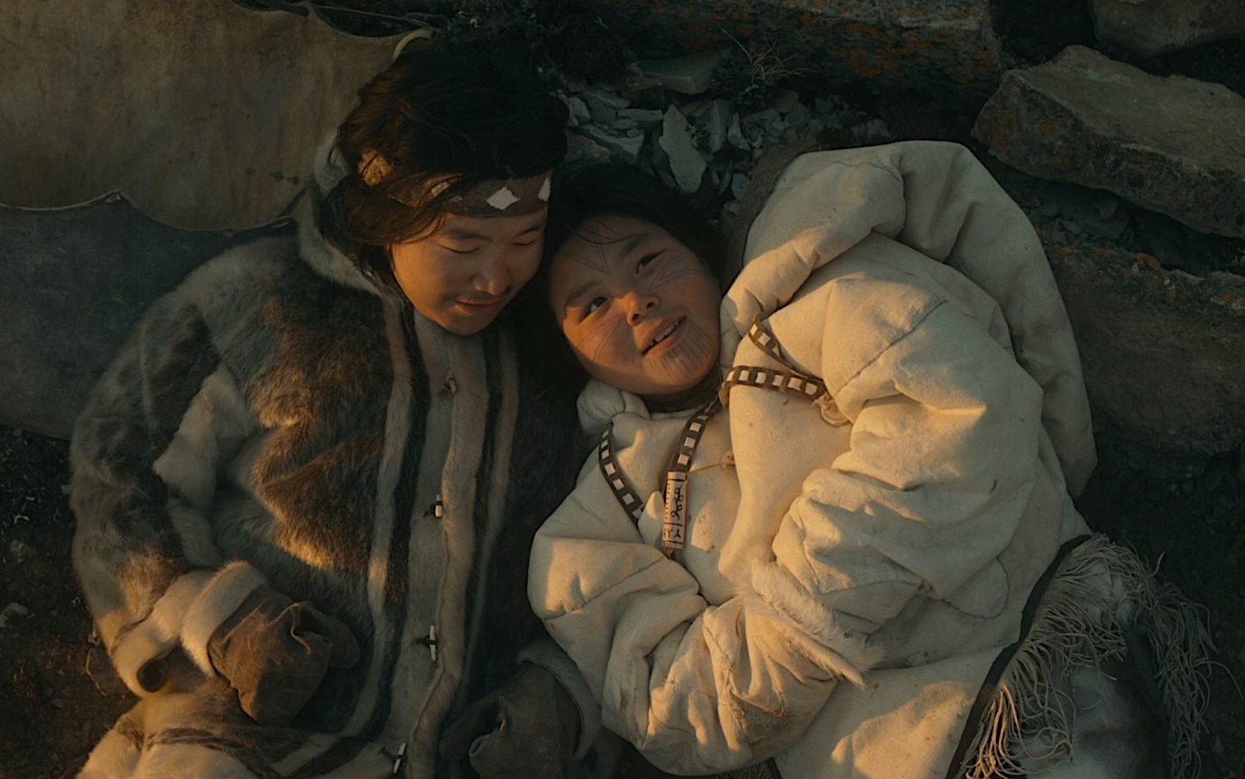 Two people in warm, fur-lined clothing lie close together on the ground among rocks, looking at each other in soft, warm light.