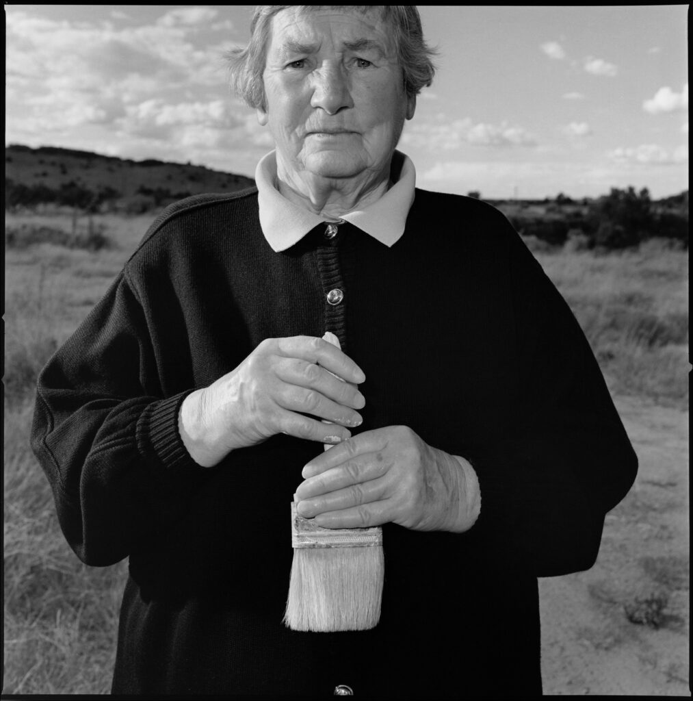 Black-and-white portrait of an older woman standing outdoors in a dry, open landscape under a cloudy sky. She faces the camera directly with a calm, serious expression. She wears a dark sweater with a light collared shirt underneath and holds a large paintbrush with both hands at chest level.