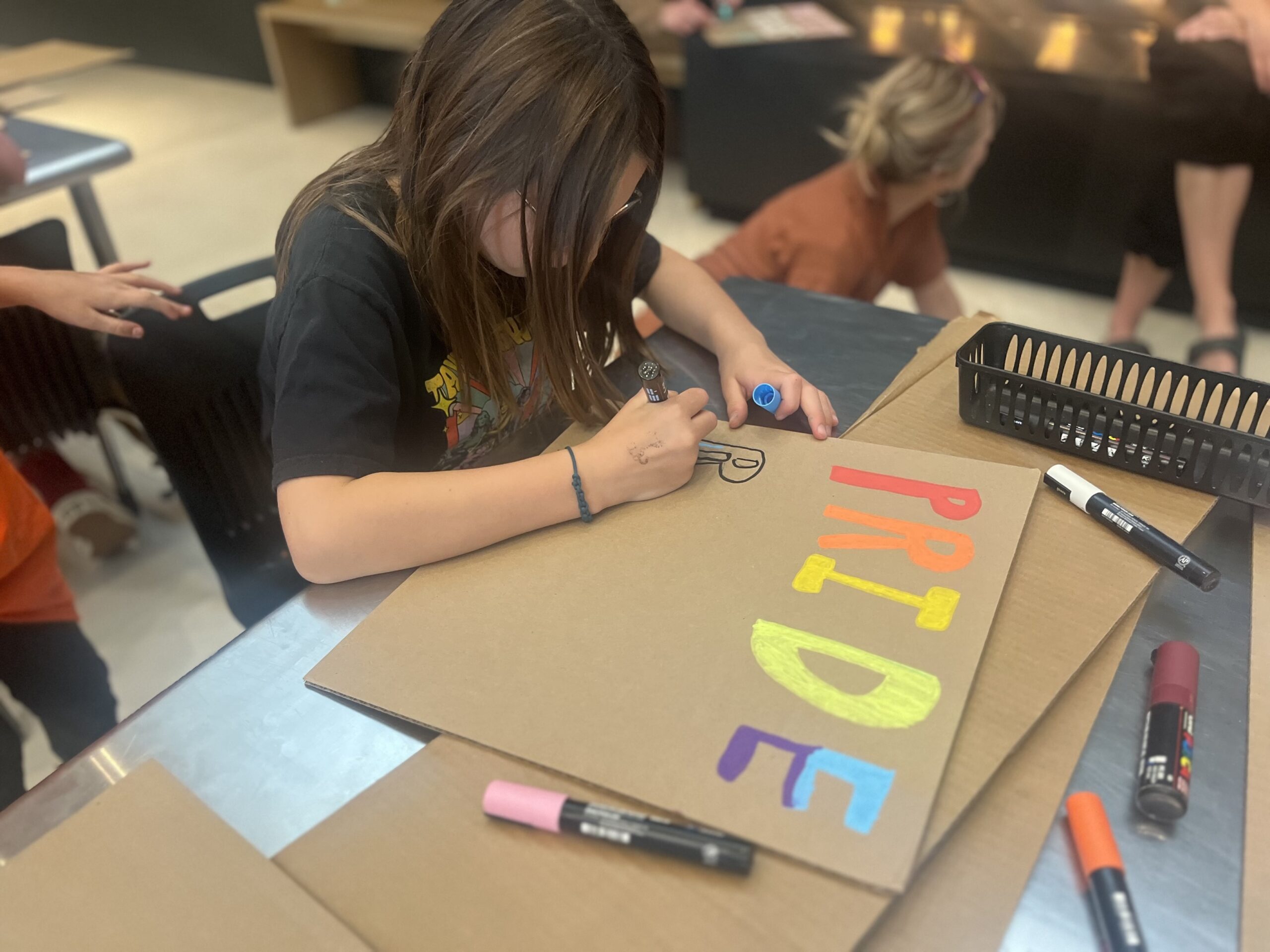 A child sits at a table creating a colorful sign on cardboard with markers. The sign spells “PRIDE” in bright rainbow colors, and art supplies are scattered around the workspace while other people work in the background.