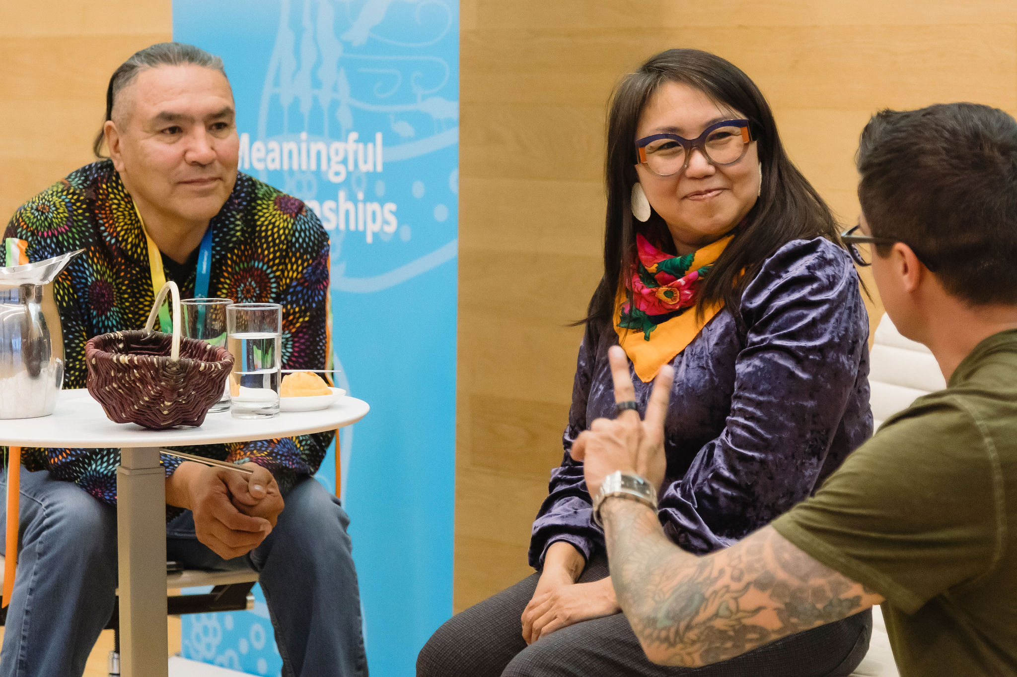 Three people participate in a seated discussion at an indoor event. On the left, a person in a brightly patterned shirt sits beside a small round table holding a water pitcher, glasses, and a woven basket. On the right, a person wearing glasses, a purple velvet top, and a colorful scarf smiles while listening to another participant in the foreground who is gesturing with their hand. A blue banner and wood-panelled wall form the background.