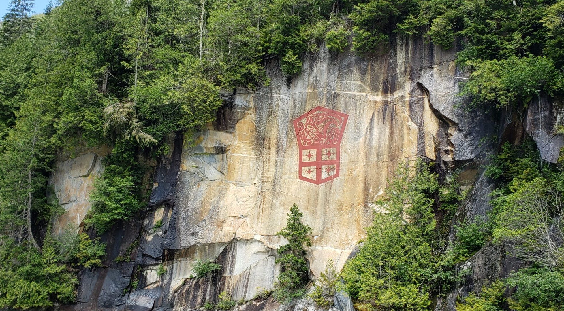 A large red design painted on a tall granite cliff face. The design shows a Northwest Coast formline creature above a shield divided into four sections, each containing a four-pointed star. Dense green coastal forest surrounds the rock.
