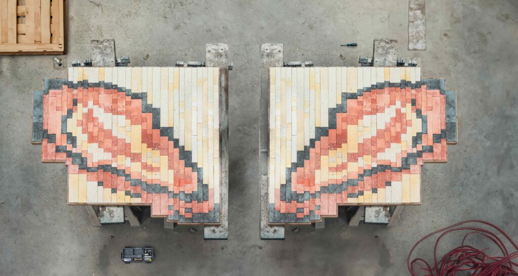 Overhead view of two rectangular tile panels resting on worktables in a workshop. Each panel features a pixelated, pink-and-cream oval pattern outlined in dark gray, resembling oysters. The surrounding concrete floor contains tools, wooden pallets, and coiled red tubing.