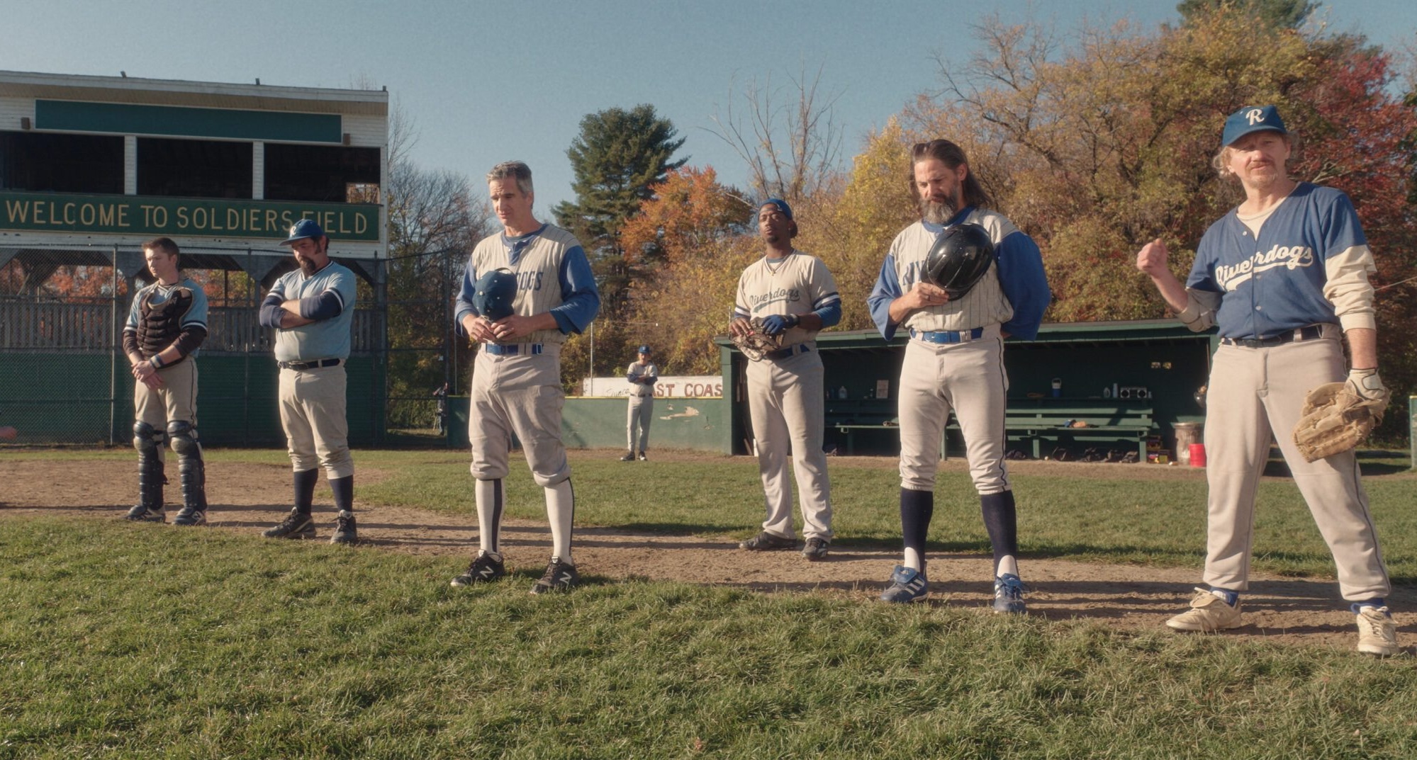 A group of adult baseball players in uniform stand on a baseball field near the dugout, listening during a break in play on a sunny autumn day.