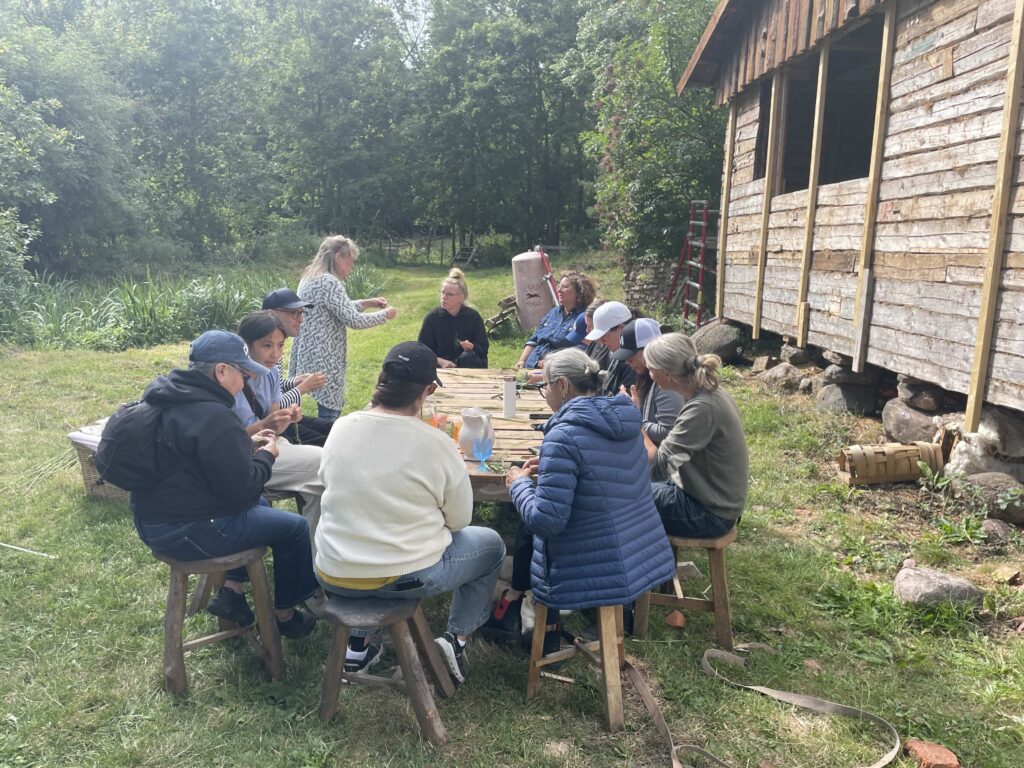 A group of people sit around a long wooden table outdoors beside a rustic timber building, participating in a hands-on workshop. One person stands and demonstrates an activity while others watch and work with materials in front of them. The gathering takes place on a grassy lawn surrounded by trees and garden plants.