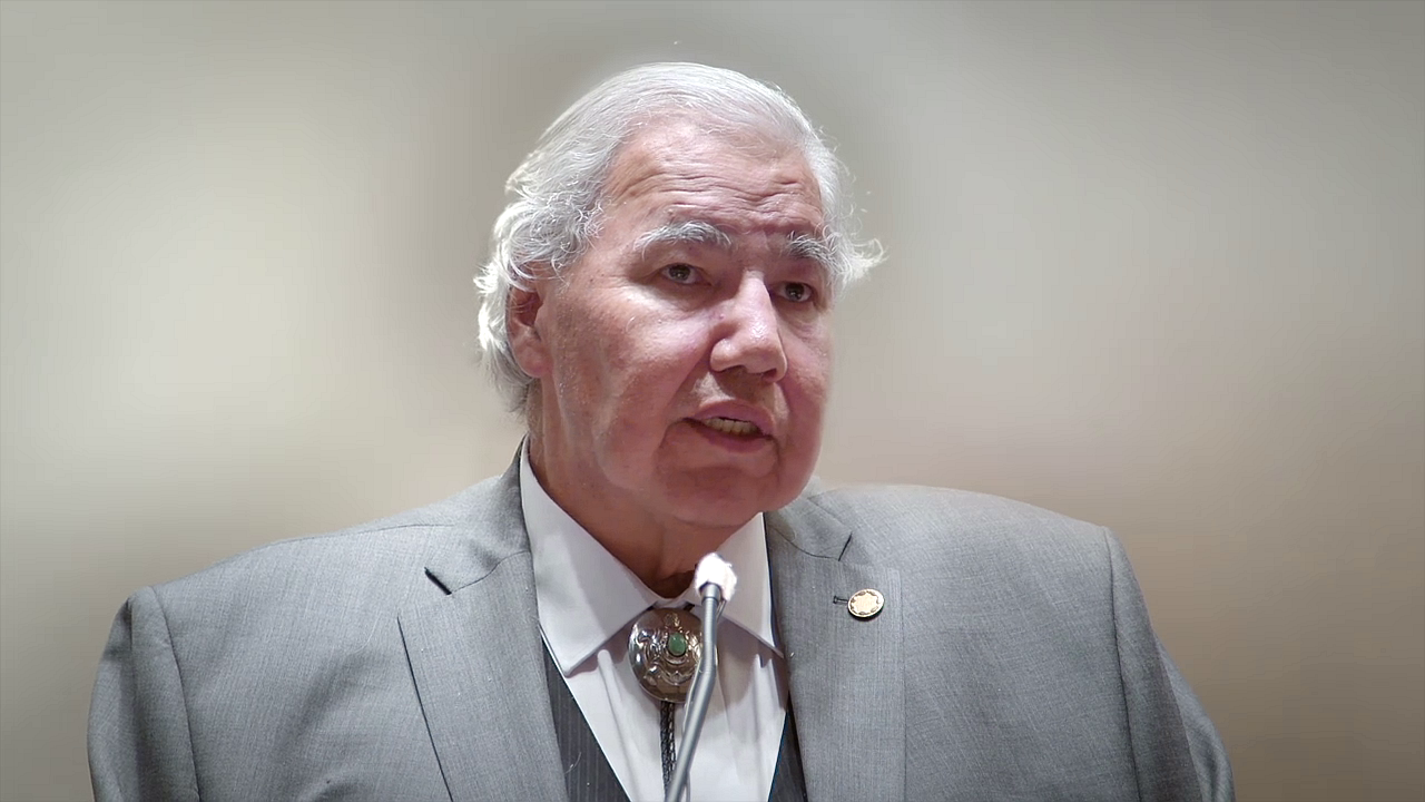 Senator Murray Sinclair speaks into a microphone while wearing a light grey suit, white shirt, and bolo tie, his expression thoughtful against a plain background.