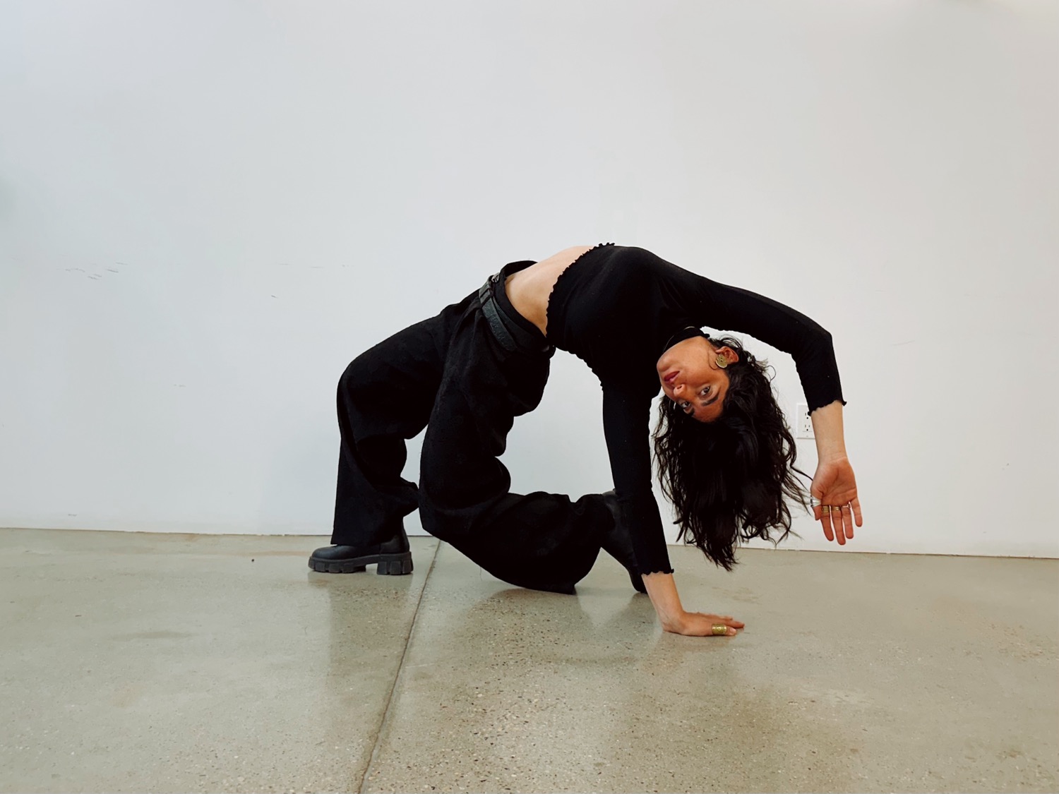 A dancer in all black arches backward into a bridge-like pose on a polished concrete floor, long dark hair sweeping toward the ground.