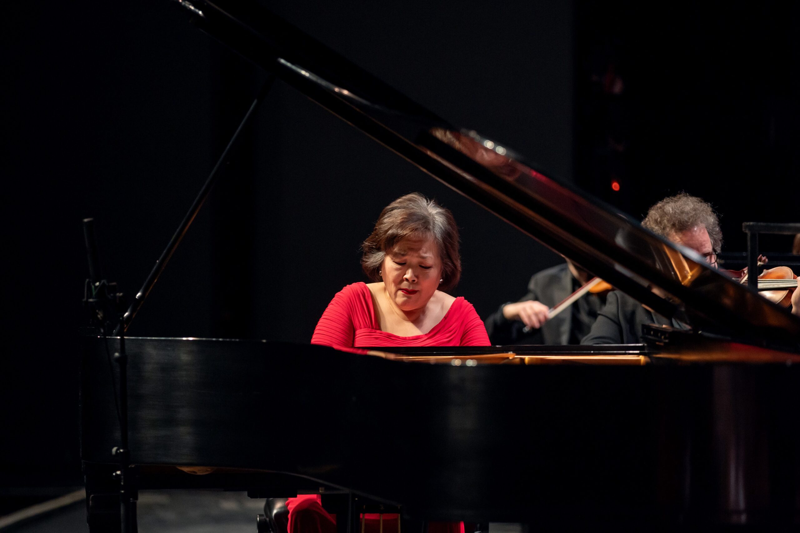 A pianist in a red dress performs at a grand piano on stage, with orchestra musicians visible in the background.