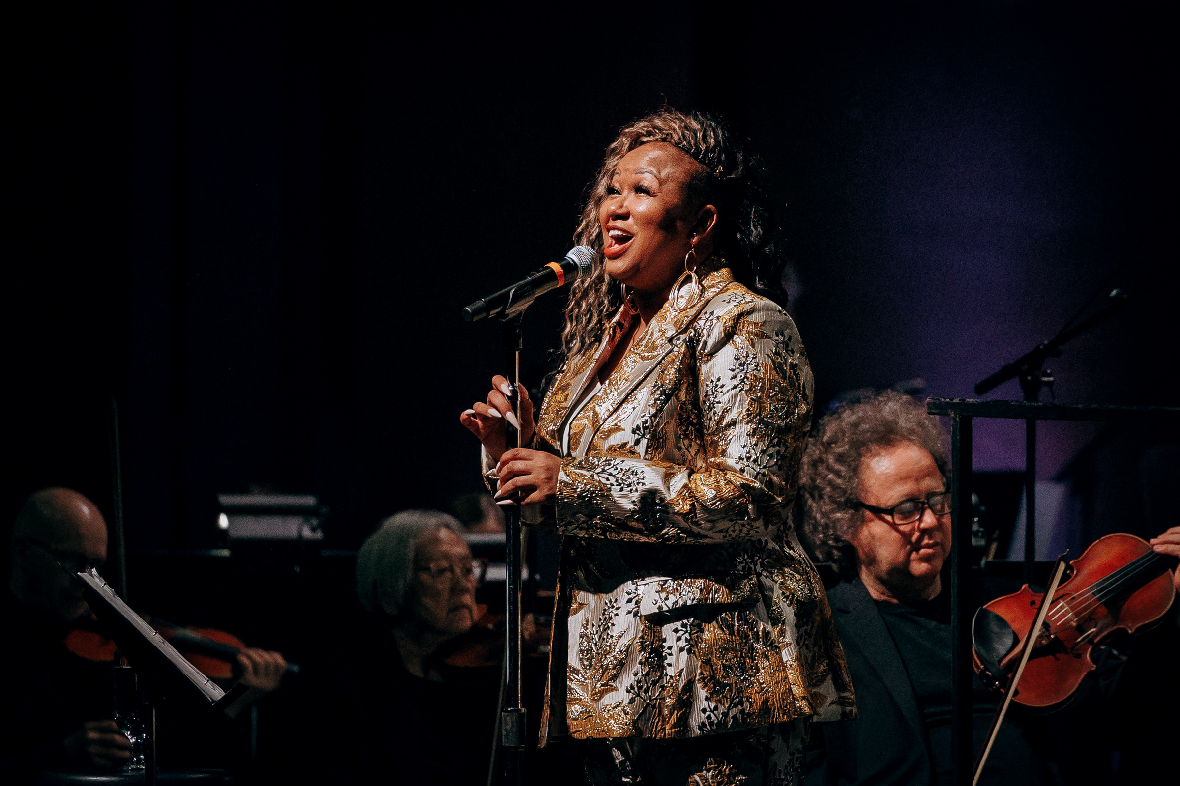 A singer in a gold-patterned jacket performs on stage with a microphone, accompanied by seated orchestra musicians playing string instruments.