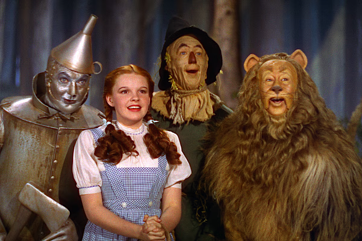 Four characters in colorful costumes stand together in a forest setting, smiling and gazing ahead, including a girl in a blue dress, a tin figure, a scarecrow, and a lion.