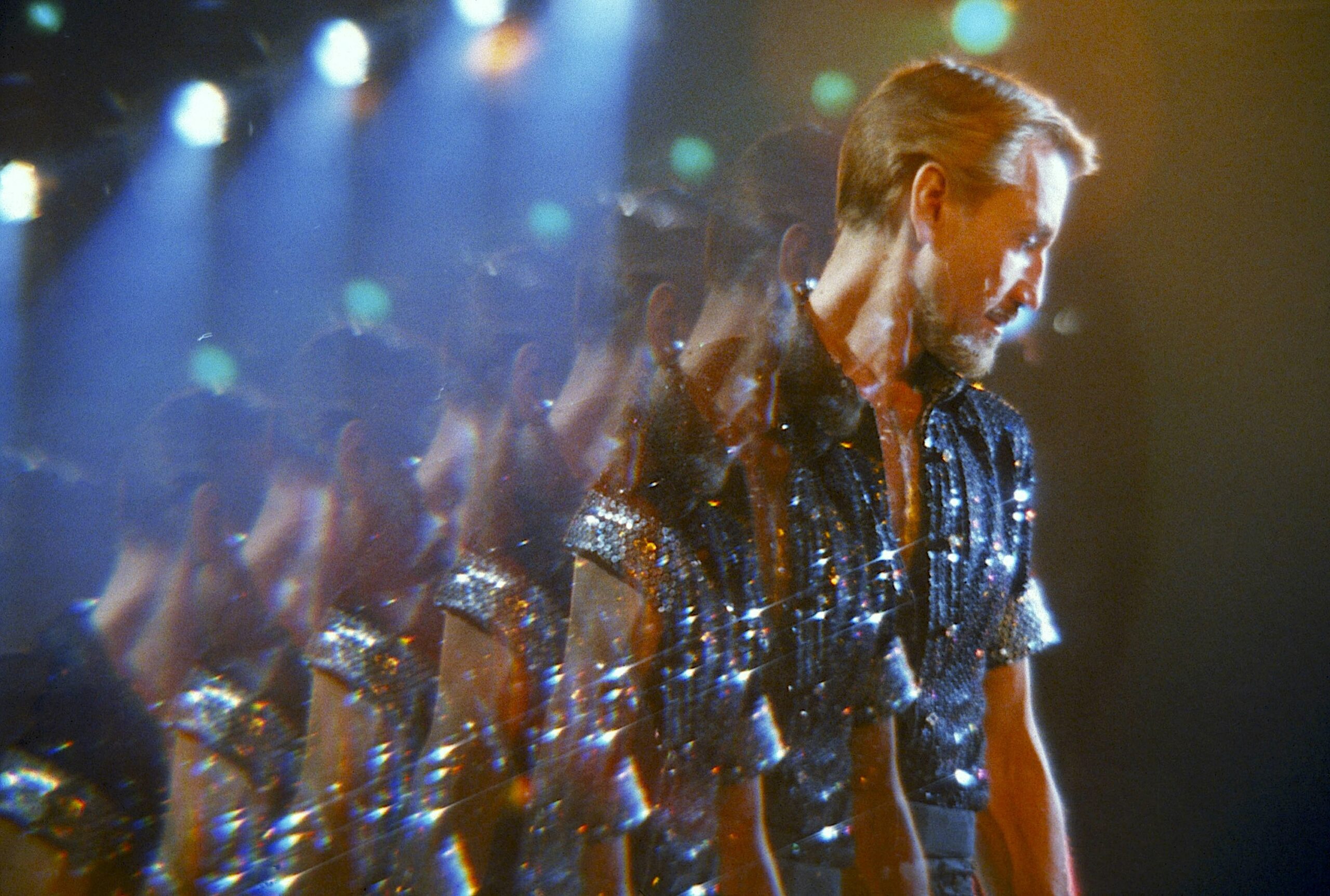 A performer in a sparkling shirt stands under stage lights, captured through a prism effect that creates multiple overlapping reflections.