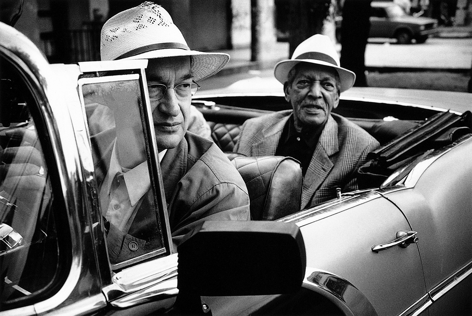 Black-and-white photo of two older men in Panama hats sitting in a vintage convertible on a city street.