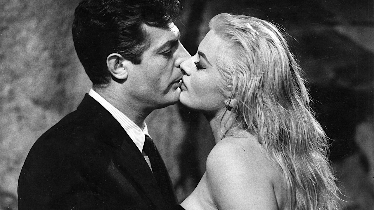 Black-and-white close-up of a man in a suit and a woman with long light hair leaning in for a kiss, their faces nearly touching in an intimate moment.