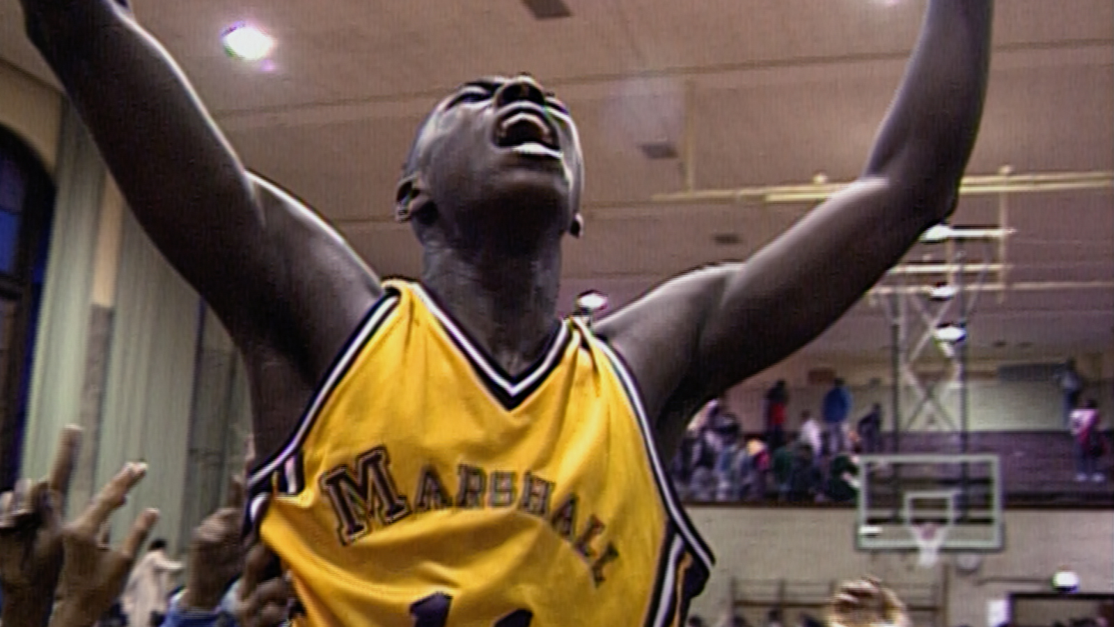 A basketball player in a yellow jersey raises both arms in celebration as a crowd surrounds him on a gym court after a game.