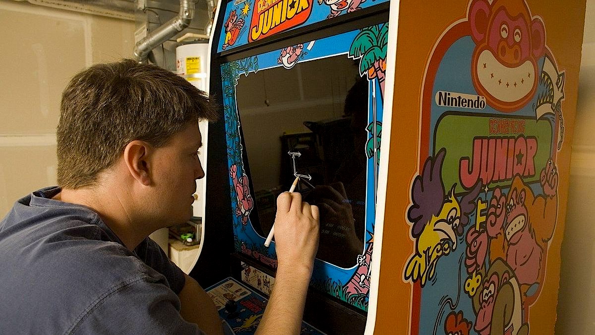 A man sits in front of a vintage arcade cabinet, carefully marking the screen while working on or examining the machine in a garage or workshop.