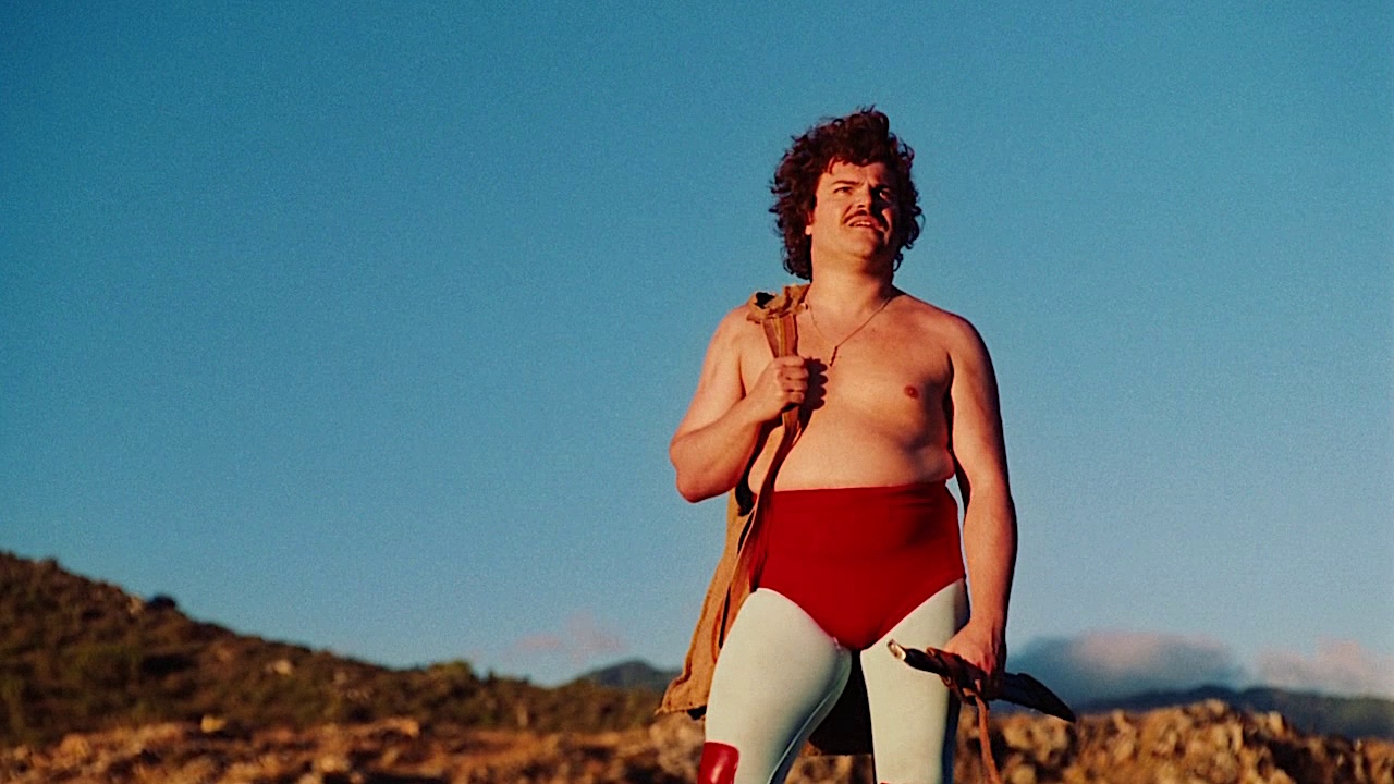 A wrestler in red trunks and white tights stands on a rocky landscape under a clear blue sky, looking off into the distance while holding a bag over one shoulder.