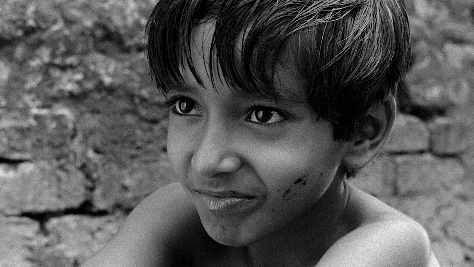 Black-and-white close-up of a young child with a faint smile and dirt smudges on one cheek, looking off-camera with a stone wall softly blurred in the background.