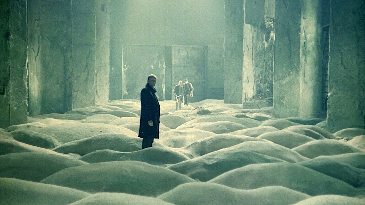 A lone figure stands in a vast, decaying industrial hall where the floor is covered in undulating white foam or snow-like mounds, while two distant figures walk through the hazy, green-tinted space.