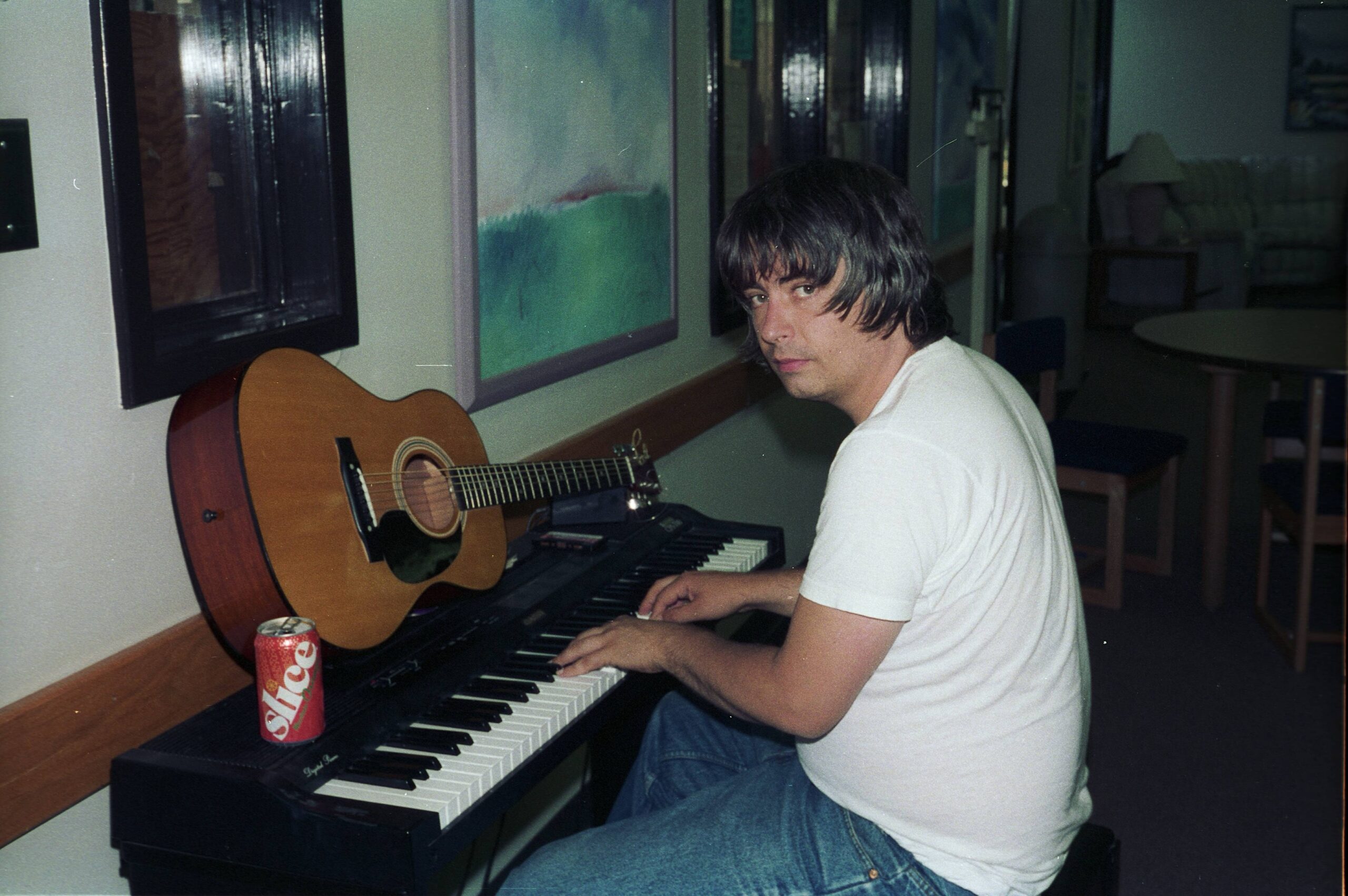 A person in a white T-shirt sits at a keyboard with an acoustic guitar beside them, looking toward the camera in a dimly lit room.