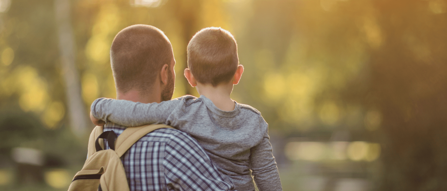An adult carries a young child on their hip while walking through a sunlit park, both seen from behind with the child's arm around the adult's shoulder."